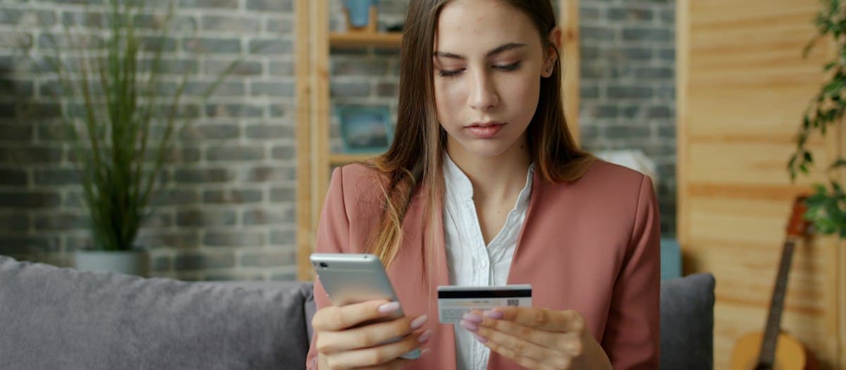 Mujer en el sillón de su casa pagando en línea con su celular y una tarjeta